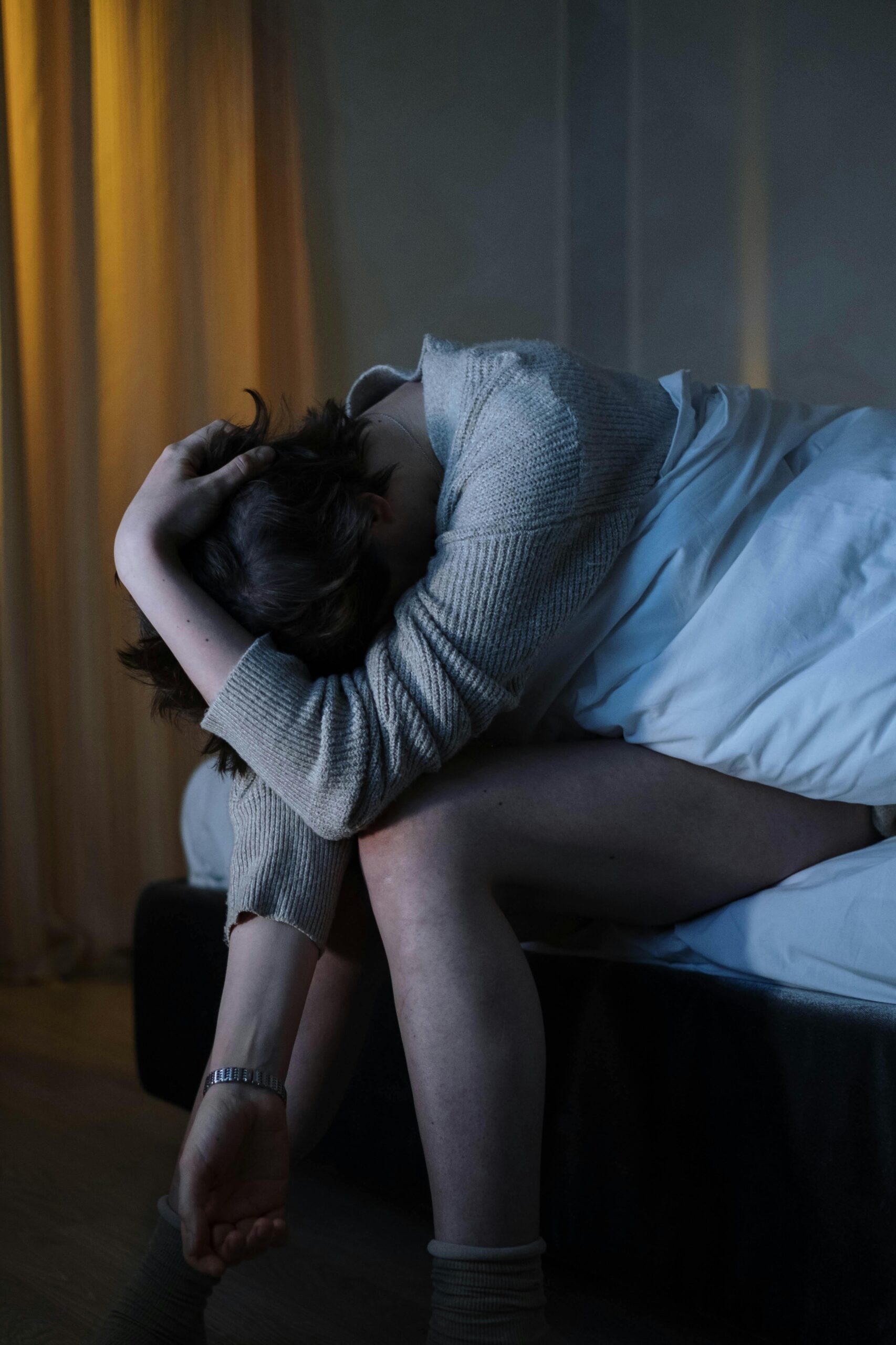 A person sitting on a bed, slumped over and clutching their head.