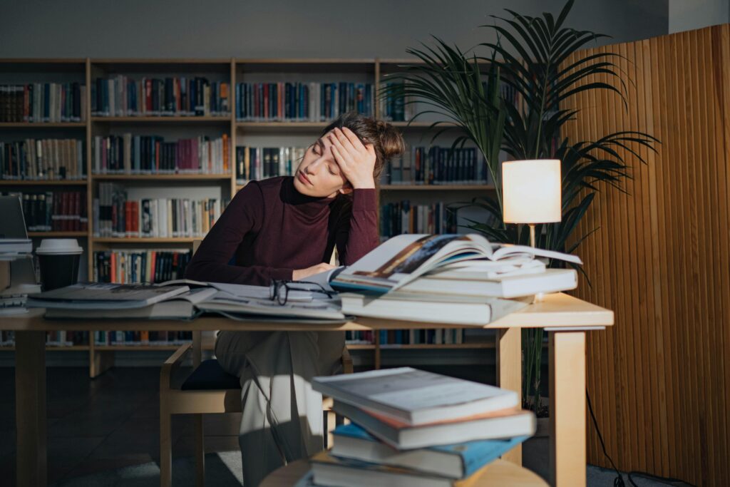A stressed woman sitting a desk overrun by books.
